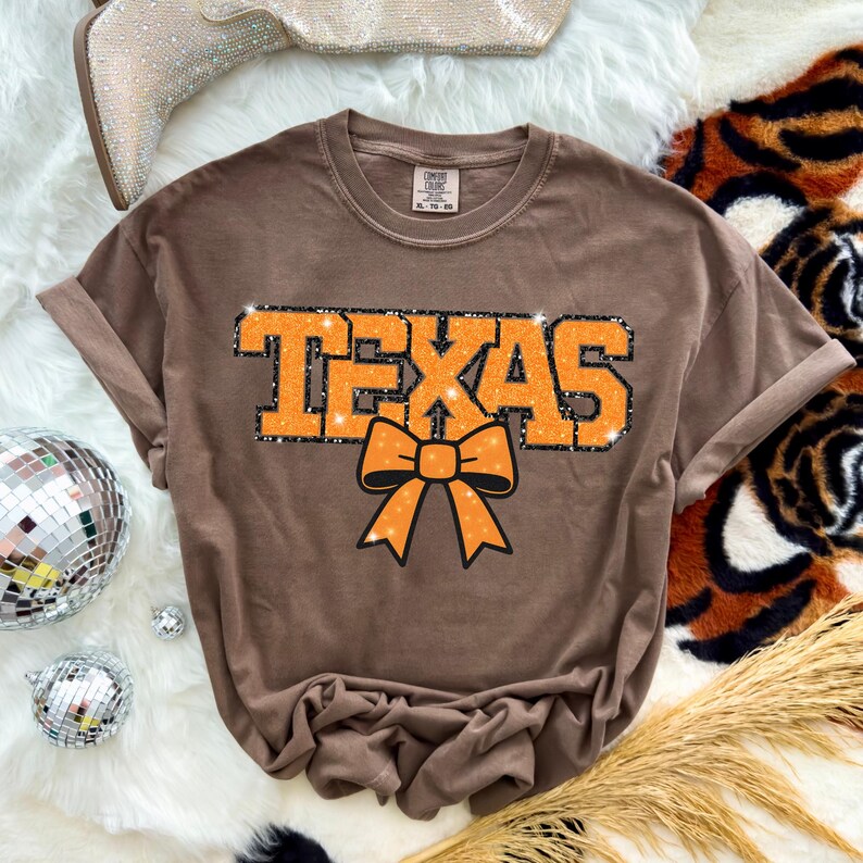 May include: A brown t-shirt featuring the word "TEXAS" in orange glitter lettering and an orange bow graphic. The shirt has rolled-up sleeves and a crew neck. The shirt is on a white surface.