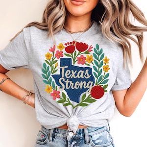 May include: A light gray t-shirt featuring a floral wreath design with the words "Texas Strong" in a blue Texas outline. The wreath includes red, yellow, and pink flowers with green leaves. The shirt is tied at the waist.