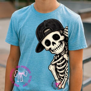 Op de afbeelding: Een gem&ecirc;leerd lichtblauw T-shirt met korte mouwen en een afbeelding van een skelet met een zwarte honkbalpet. Het skelet staat aan de rechterkant van het shirt en kijkt naar links. Een roze striksymbool en tekst zijn zichtbaar op het shirt.