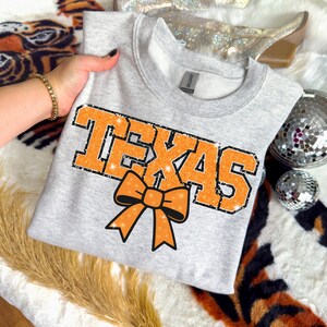 May include: A light grey sweatshirt featuring the word "TEXAS" in orange block letters with a black glitter outline. An orange bow graphic is below the word. The sweatshirt is folded.