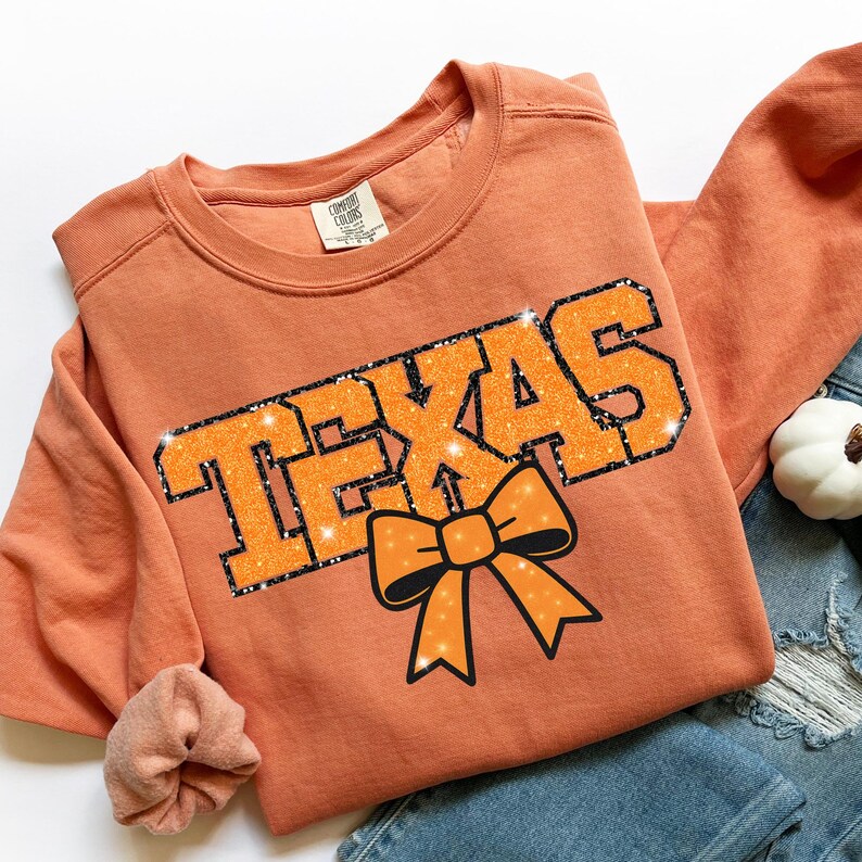 May include: Orange sweatshirt featuring the word "TEXAS" in glittery orange letters with a black outline. A matching orange bow is below the word. The sweatshirt is folded on top of blue jeans.