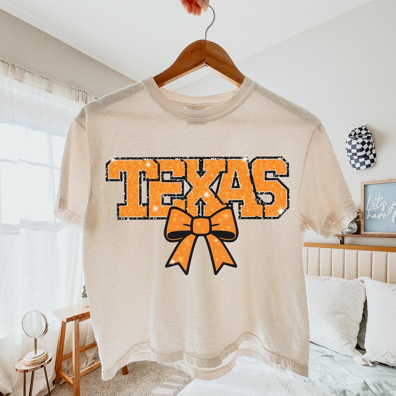 May include: Cream-coloured t-shirt featuring the word "TEXAS" in orange glitter lettering and an orange bow graphic. The shirt is displayed on a wooden hanger.