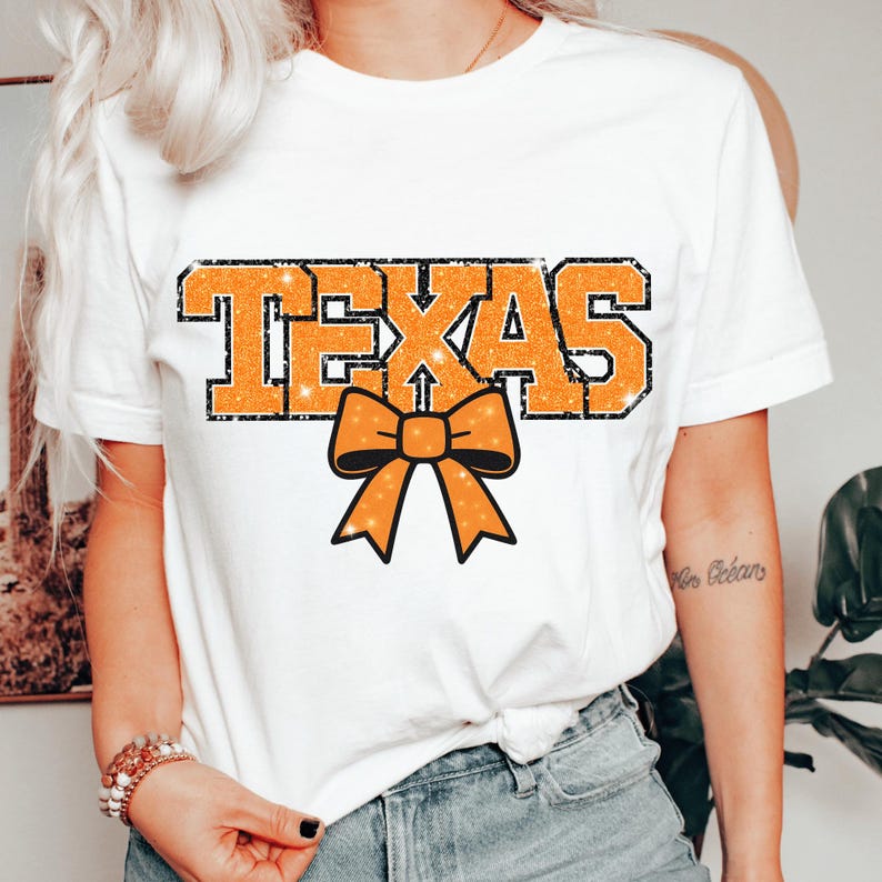 May include: White t-shirt with the word "TEXAS" in orange glitter lettering and a black outline. An orange glitter bow is below the text. The shirt is a casual style.