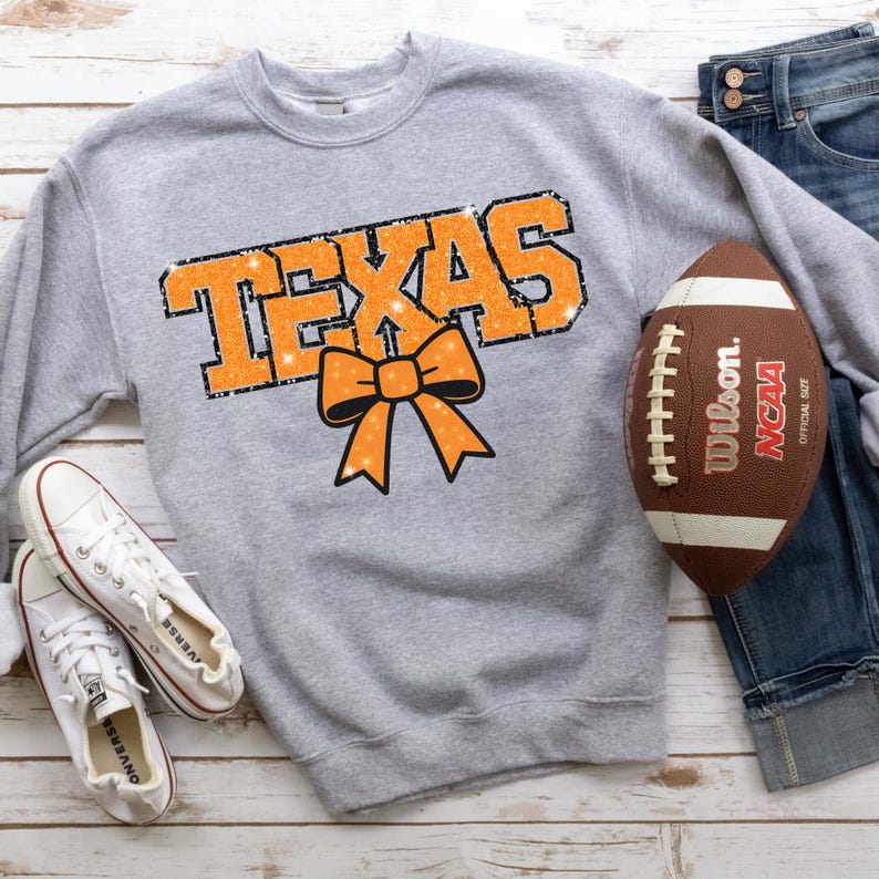 May include: A grey sweatshirt featuring the word "TEXAS" in orange glitter lettering and an orange bow graphic. A brown and white American football and a pair of white Converse trainers are also in the image. Blue jeans are visible.