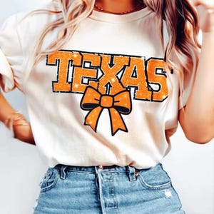 May include: Cream-coloured t-shirt featuring the word "TEXAS" in orange glitter lettering and a matching bow graphic. The shirt is paired with blue denim shorts. The design is centered on the front of the shirt.