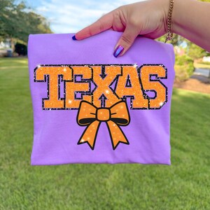 May include: A lilac t-shirt with the word "TEXAS" in orange glitter lettering and a black outline. An orange bow with black accents is below the text. The shirt is held up against a green grassy background.