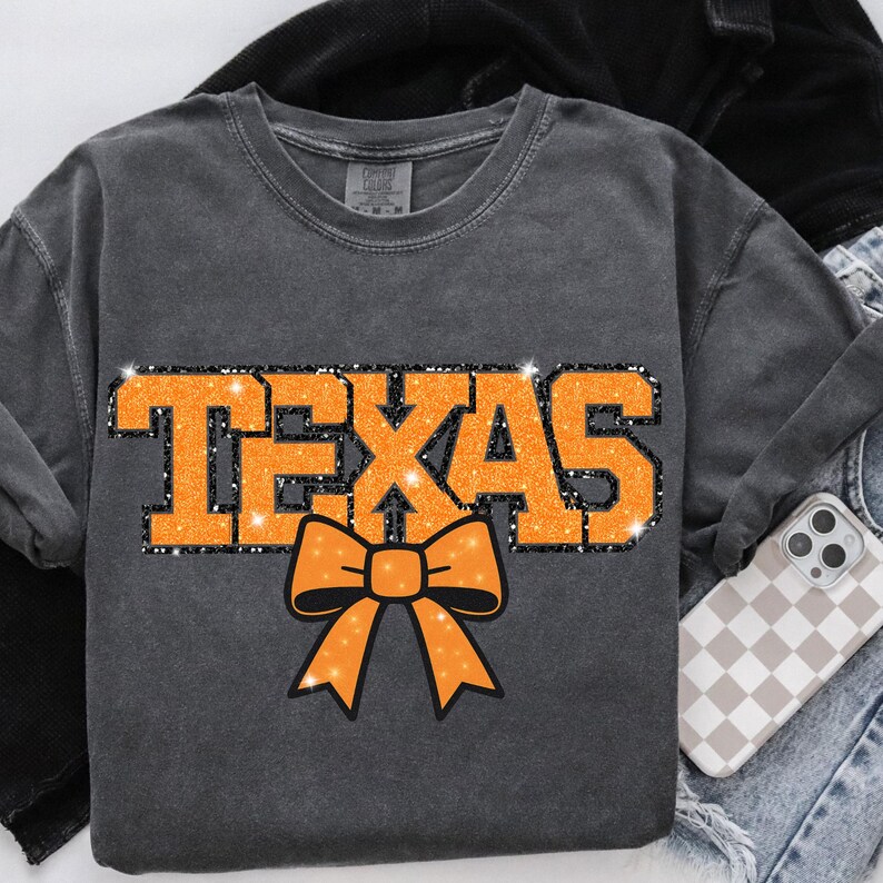 May include: A grey long-sleeved shirt featuring the word "TEXAS" in orange glitter lettering. Beneath the word is an orange bow. The shirt is on a white surface with a black jacket, jeans, and a phone case in the background.