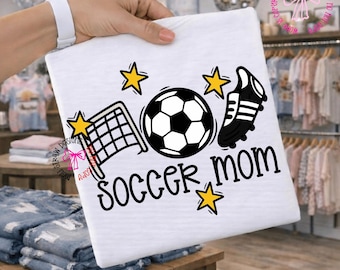 Könnte beinhalten: Weißes T-Shirt mit einem schwarz-weißen Fußball, einem Fußballtor und einem Fußballschuh-Motiv. Der Text "Soccer Mom" ist in Schwarz gedruckt, mit gelben Sternakzenten. Das Shirt wird von einer Person gehalten.