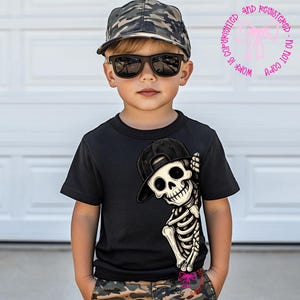 Op de afbeelding: Een zwart T-shirt met korte mouwen en een afbeelding van een skelet met een zwarte honkbalpet. Het skelet bevindt zich aan de rechterkant van het T-shirt. Het T-shirt wordt gedragen met een camouflage honkbalpet en een zwarte zonnebril.
