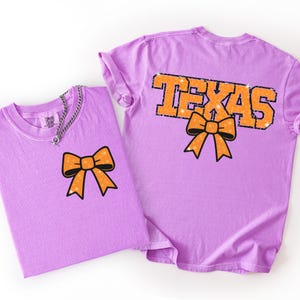 May include: Two lilac-coloured t-shirts. One shirt has an orange bow graphic on the front. The other shirt has the word "TEXAS" in orange with a black outline and an orange bow graphic. Both shirts are made of a soft material.