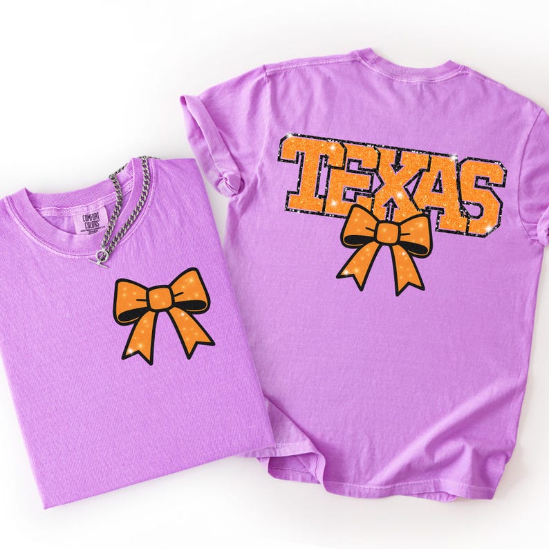 May include: Two lilac-coloured t-shirts. One shirt has an orange bow graphic on the front. The other shirt has the word "TEXAS" in orange with a black outline and an orange bow graphic. Both shirts are made of a soft material.
