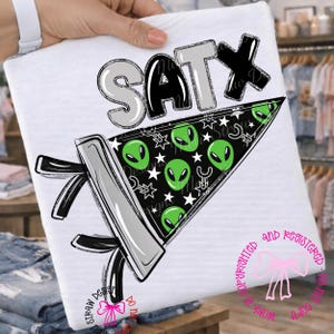 May include: A white t-shirt featuring a graphic design with the text SATX in silver and black. Below the text is a triangular pennant in black with a pattern of green alien faces, white stars, and small silver slingshots, accented with black ribbons.