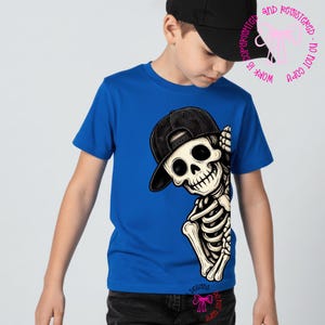 Op de afbeelding: Een felblauw t-shirt met korte mouwen en een grafische print van een skelet met een zwarte honkbalpet. Het skelet is aan de rechterkant van het shirt geplaatst en lijkt met zijn ribbenkast en arm vanaf de zijkant naar buiten te kijken.