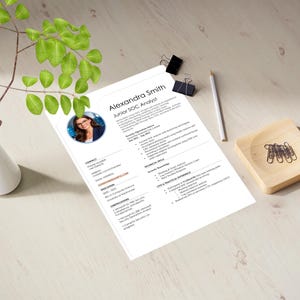 May include: A resume for Alexandra Smith, a Junior SOC Analyst, is displayed on a light wooden surface. The resume includes a photo, contact information, and details about education, skills, and experience. A white vase with green leaves, a pencil, paper clips, and a wooden dish are also present.