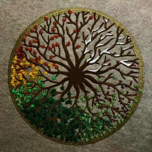 Puede incluir: Dise&ntilde;o circular bordado de un &aacute;rbol con ramas de diferentes colores que representan las cuatro estaciones. El &aacute;rbol es marr&oacute;n con hojas verdes, amarillas, rojas y blancas.