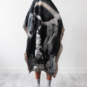 May include: A woven throw blanket featuring a dark, monochromatic artistic print of a human figure and abstract shapes. The rectangular blanket has a fringed edge in a light tan color and is draped over a form, revealing only legs in gray socks.