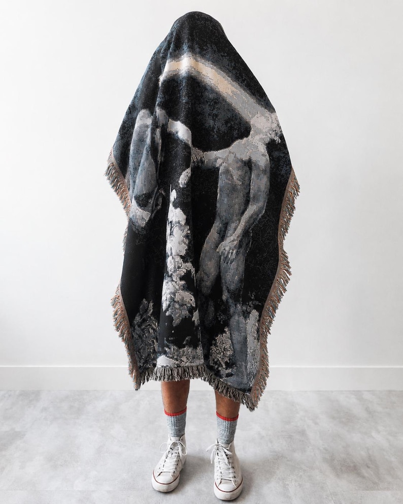 May include: A woven throw blanket featuring a dark, monochromatic artistic print of a human figure and abstract shapes. The rectangular blanket has a fringed edge in a light tan color and is draped over a form, revealing only legs in gray socks.