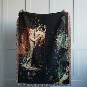 May include: A rectangular woven throw blanket with a black background featuring a detailed tapestry-style illustration of a figure holding a large ornate vessel. The design includes warm copper, gold, and deep green tones with a fringed edge.