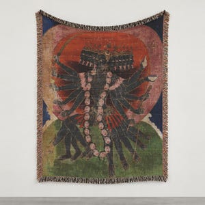 May include: A woven throw blanket featuring a multi-armed, dark-skinned deity with multiple heads, wearing a garland of skulls. The figure is set against a large red circular halo, a blue sky, and a green landscape, finished with fringed edges.