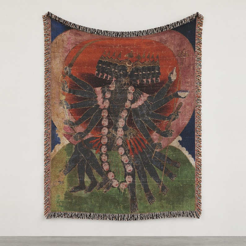 May include: A woven throw blanket featuring a multi-armed, dark-skinned deity with multiple heads, wearing a garland of skulls. The figure is set against a large red circular halo, a blue sky, and a green landscape, finished with fringed edges.