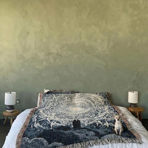 May include: A woven throw blanket featuring a dark blue and white celestial graphic of a swirling galaxy and two figures standing on a hill. The blanket has a fringed edge and is draped over a white bedspread with a small tan dog sitting on top.