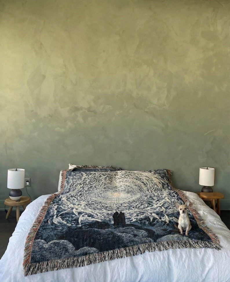 May include: A woven throw blanket featuring a dark blue and white celestial graphic of a swirling galaxy and two figures standing on a hill. The blanket has a fringed edge and is draped over a white bedspread with a small tan dog sitting on top.