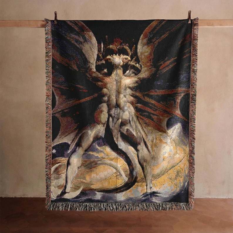 Puede incluir: Una manta tejida rectangular que presenta una ilustraci&oacute;n oscura y detallada de una criatura alada y musculosa con una cola larga. El dise&ntilde;o utiliza tonos negros, dorados y marrones, con un borde decorativo de flecos arriba y abajo.