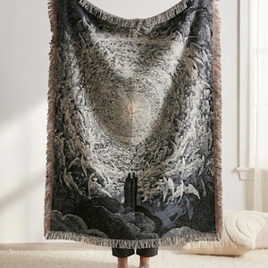 May include: A rectangular woven throw blanket featuring a detailed monochromatic illustration of a celestial scene with a central radiant light source, winged figures, and two dark silhouettes standing at the base, finished with fringed edges.
