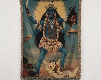 Puede incluir: Una manta de tapiz tejida que presenta una figura de piel azul con cuatro brazos, que lleva una corona y una guirnalda de calaveras. La figura sostiene una espada y una cabeza cortada sobre un cuenco, de pie sobre una figura reclinada.