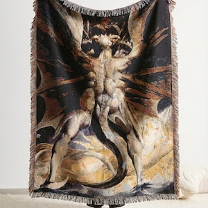 Puede incluir: Una manta tejida rectangular que presenta una ilustraci&oacute;n detallada de una criatura alada y con cuernos con una cola larga. El dise&ntilde;o utiliza un fondo negro oscuro con tonos beige, marrones y dorados. La manta tiene bordes con flecos.