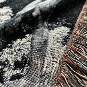 May include: A woven throw blanket featuring a high-contrast black and white abstract pattern with a textured surface. The edge of the blanket is finished with a decorative fringe containing strands of black, white, tan, and muted blue threads.