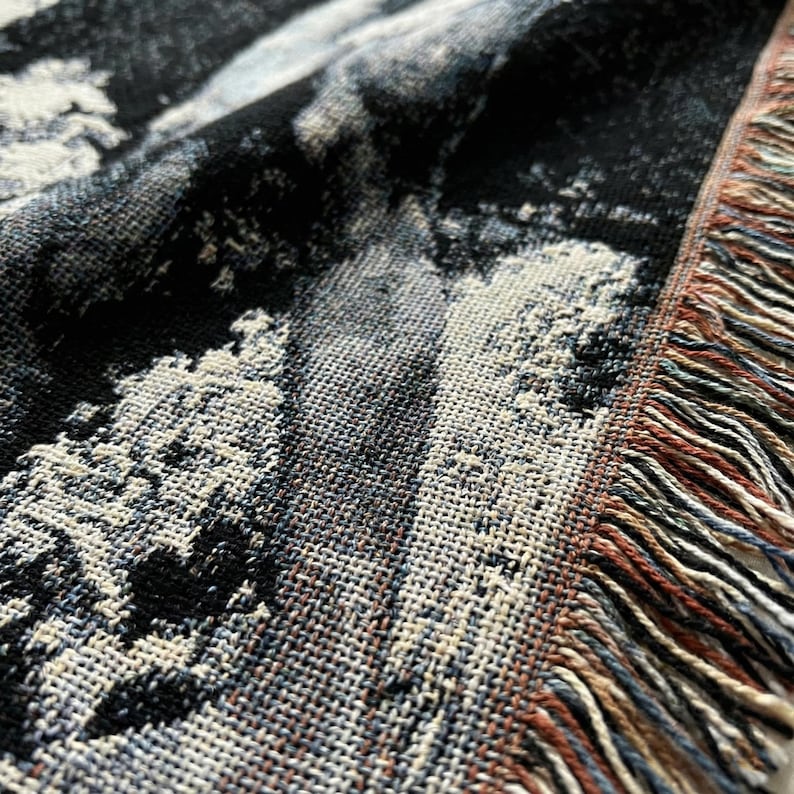 May include: A woven throw blanket featuring a high-contrast black and white abstract pattern with a textured surface. The edge of the blanket is finished with a decorative fringe containing strands of black, white, tan, and muted blue threads.