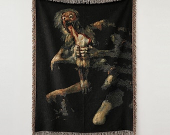 Puede incluir: Una manta tejida con una reproducción de Saturno devorando a su hijo de Francisco Goya. El tapiz oscuro y texturizado muestra una figura de ojos muy abiertos consumiendo una forma más pequeña sobre un fondo negro, con flecos.