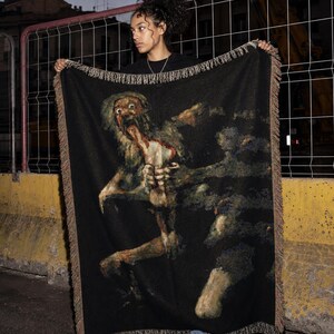 Pu&ograve; includere: Una coperta rettangolare tessuta che presenta una rappresentazione scura e dettagliata del dipinto Saturno che divora il figlio. La coperta ha uno sfondo nero con un bordo sfrangiato in toni terra tenui, raffigurante una figura con gli occhi sgranati.