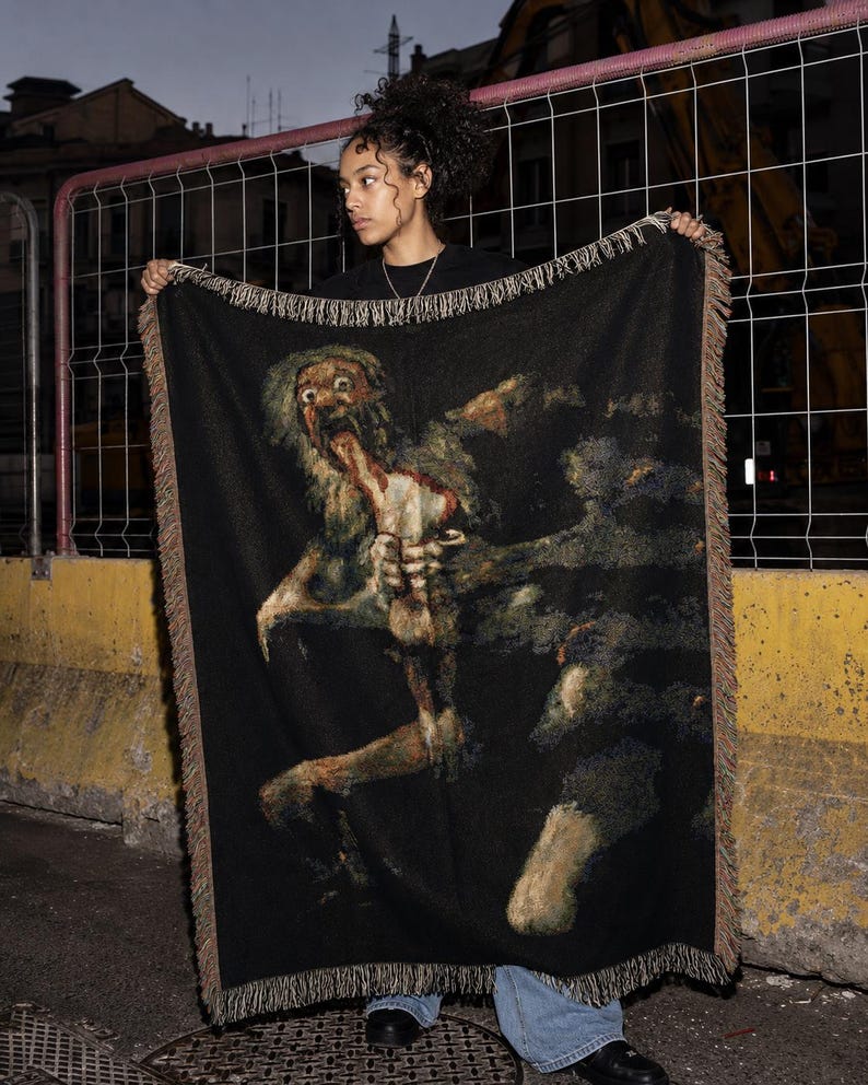 Pu&ograve; includere: Una coperta rettangolare tessuta che presenta una rappresentazione scura e dettagliata del dipinto Saturno che divora il figlio. La coperta ha uno sfondo nero con un bordo sfrangiato in toni terra tenui, raffigurante una figura con gli occhi sgranati.