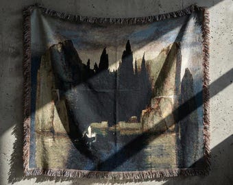 May include: A woven throw blanket featuring a dark, moody landscape scene with towering rock formations, deep blue water, and a dramatic sky. The rectangular textile has fringed edges on all four sides and a detailed, textured appearance.