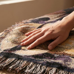 Puede incluir: Una manta tejida con un patr&oacute;n abstracto multicolor en tonos morado, negro, bronceado y dorado. El tejido tiene una textura similar a un tapiz y est&aacute; rematado con un fleco decorativo a lo largo del borde curvo.