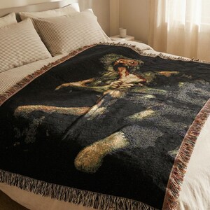 Pu&ograve; includere: Una coperta in tessuto nero con una dettagliata rappresentazione artistica multicolore di una figura con un'espressione dagli occhi sgranati. La coperta rettangolare presenta una superficie testurizzata e bordi con frange.