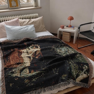 May include: A rectangular woven throw blanket featuring a dark black background with a detailed tapestry-style illustration of a classical figure holding a vessel, accented with green, brown, and cream tones and finished with a fringed edge.