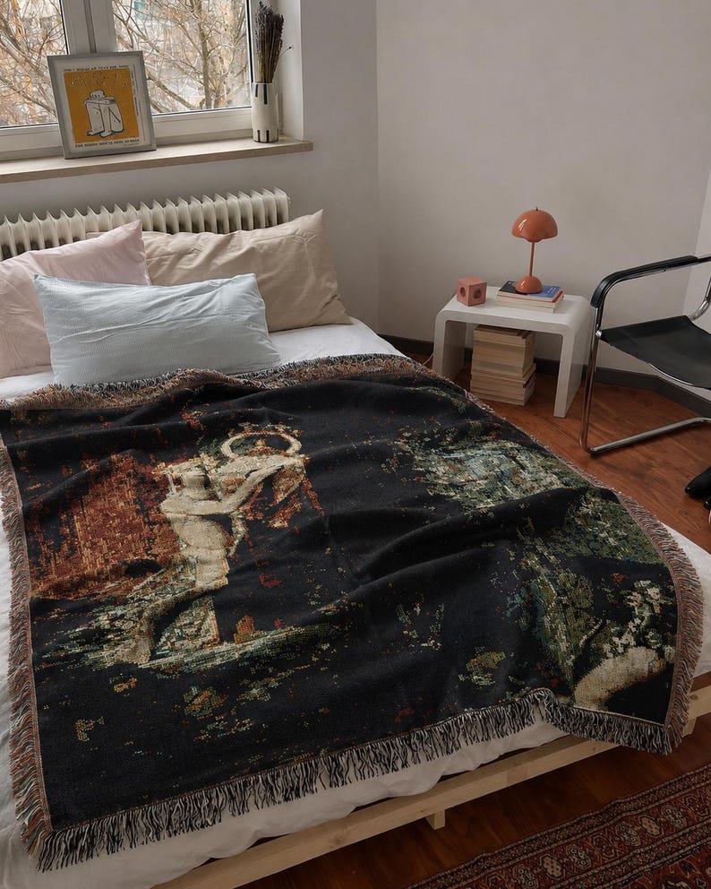 May include: A rectangular woven throw blanket featuring a dark black background with a detailed tapestry-style illustration of a classical figure holding a vessel, accented with green, brown, and cream tones and finished with a fringed edge.