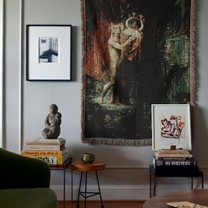 May include: A large rectangular woven wall tapestry featuring a dark, textured scene of a figure holding a lyre, displayed alongside framed art prints, stacks of books, a small stone sculpture, and a small green ceramic bowl.