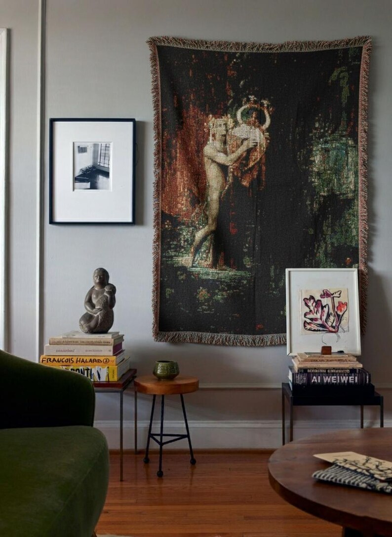 May include: A large rectangular woven wall tapestry featuring a dark, textured scene of a figure holding a lyre, displayed alongside framed art prints, stacks of books, a small stone sculpture, and a small green ceramic bowl.