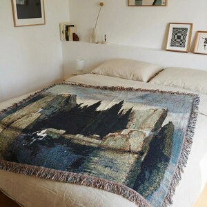 Puede incluir: Una manta tejida rectangular que presenta una reproducci&oacute;n detallada de la pintura La isla de los muertos de Arnold B&ouml;cklin. El tejido muestra cipreses oscuros, acantilados rocosos y agua azul tranquila con flecos en los cuatro lados.
