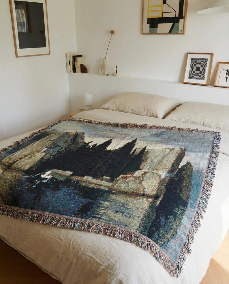 Puede incluir: Una manta tejida rectangular que presenta una reproducci&oacute;n detallada de la pintura La isla de los muertos de Arnold B&ouml;cklin. El tejido muestra cipreses oscuros, acantilados rocosos y agua azul tranquila con flecos en los cuatro lados.