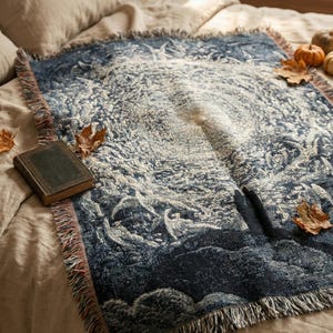 May include: A rectangular woven throw blanket featuring a dark blue and cream intricate celestial pattern with a central sunburst design, finished with fringed edges on all sides, displayed on a neutral-coloured bedspread.