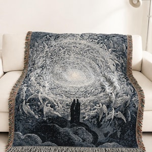 May include: A woven throw blanket featuring a dark blue and white tapestry design of two figures standing on a rocky cliff looking towards a bright, swirling celestial light surrounded by winged forms, finished with a fringed edge.