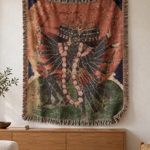 May include: A woven throw blanket featuring a multi-armed dark-skinned deity with multiple heads, adorned with a long pink floral garland. The artwork is set against a large circular orange sun, with green and blue accents and a fringed edge.