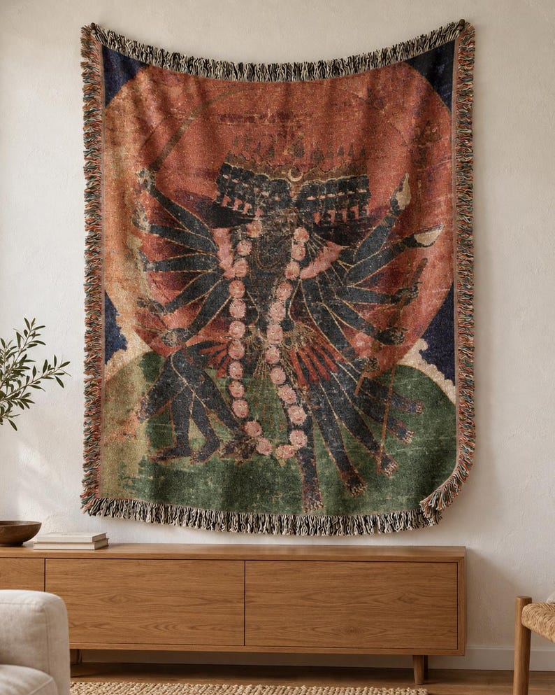 May include: A woven throw blanket featuring a multi-armed dark-skinned deity with multiple heads, adorned with a long pink floral garland. The artwork is set against a large circular orange sun, with green and blue accents and a fringed edge.
