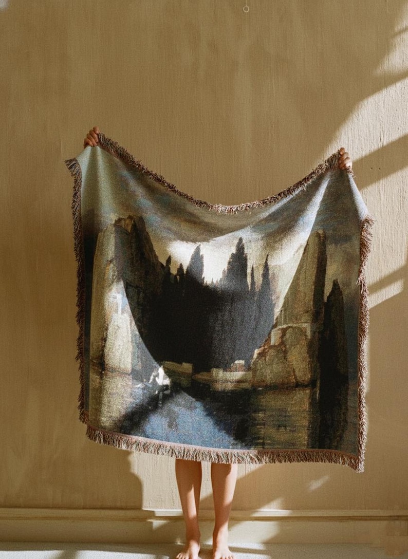 Puede incluir: Una manta tejida rectangular que presenta un paisaje oscuro y atmosf&eacute;rico con formaciones rocosas imponentes, sombras profundas y un cuerpo de agua reflectante. La manta tiene un borde con flecos y una paleta de colores apagados en azul, marr&oacute;n y gris.