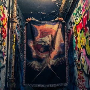 Puede incluir: Una manta tejida rectangular con un fondo oscuro y un dise&ntilde;o abstracto central en tonos naranja, amarillo y morado. La manta tiene un borde con flecos en todos sus lados y presenta una apariencia texturizada similar a un tapiz.